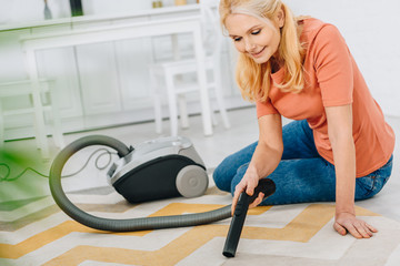 Smiling blonde woman sitting on carpet and using vacuum cleaner