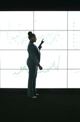 Silhouette of a black african businesswoman giving a presentation at a business conference. Pointing at a large video screen with charts and graphs next to her in a dark room