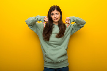 Teenager girl with green sweatshirt on yellow background showing thumb down with both hands