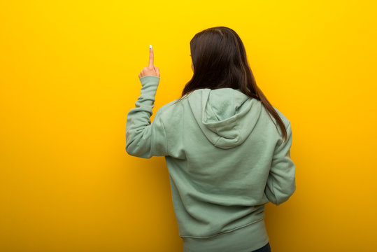 Teenager Girl With Green Sweatshirt On Yellow Background Pointing Back With The Index Finger