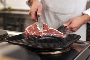 Image of european male chief in white uniform, grilling meat on at kitchen in restaurant