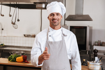 Image of positive male chief in white uniform smiling, while standing at kitchen in restaurant