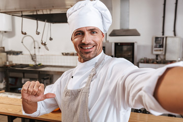 Photo of smiling male chief in apron taking selfie, while standing at kitchen in restaurant
