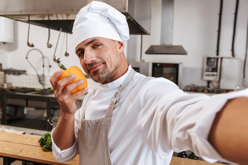 Photo of pleased male chief in apron taking selfie, while standing at kitchen in restaurant