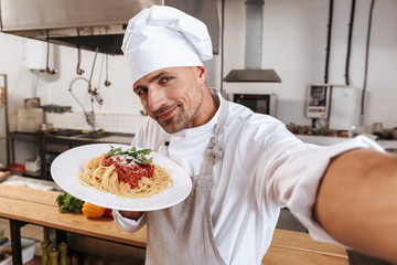 Image of handsome man chief in apron taking selfie, while standing at kitchen in restaurant