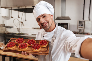Photo of cheerful male chief in white uniform taking selfie and holding plate with cakes, while cooking at kitchen in restaurant