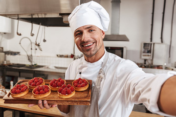 Photo of handsome male chief in white uniform taking selfie and holding plate with cakes, while cooking at kitchen in restaurant