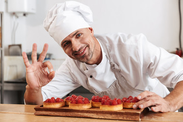 Photo of smiling male chief in white uniform holding plate with cakes, while cooking at kitchen in...