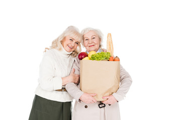 cheerful retired woman hugging friend with paper bag isolated on white