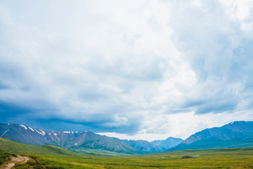 Fototapeta premium Spectacular view of distant giant mountains. Footpath through valley in highlands. Hiking path. Wonderful huge mountain range under cloudy sky. Amazing dramatic green landscape of majestic nature.