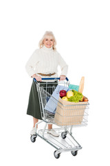 happy senior woman holding shopping cart with shopping bags and paper bag with groceries isolated on white