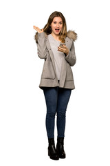 A full-length shot of a Teenager girl with coat surprised while sending a message with the mobile on isolated white background