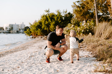 Fototapeta na wymiar Young Father and His Little Boy Son Walking and Enjoying the Nice Outdoor Weather on the Sandy Beach next to the Ocean Bay with Sail Boats in the Background