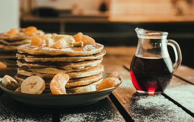 Stack of golden pancakes with bananas and oranges on wooden board covered with caster sugar. Heap of american pancakes with a jug full of syrup in kitchen.