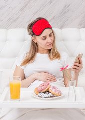 Pregnant woman using smartphone and having breakfast in bed at home