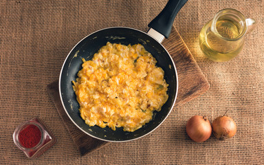 Photo of scrambled eggs with ground paprika, onions and oil from above - Rural style. Yellow scrambled eggs in black pan on jute background.