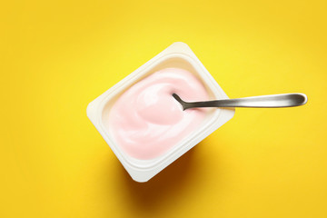 Plastic cup with creamy yogurt and spoon on color background, top view