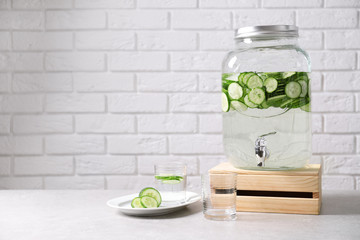 Jar dispenser of fresh cucumber water and glasses on table against brick wall. Space for text