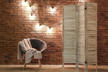 Armchair and folding screen near brick wall decorated with fairy lights