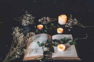Open vintage poetry book on black table surface with white lit burning candles and dried flowers....