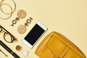 Top view of earrings, glasses, eye shadow, smartphone, mascara, bracelets, cosmetic brush and bag on yellow background