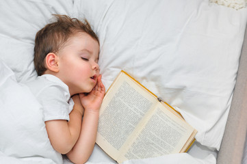 A cute toddler boy is sleeping with an old book under a white blanket.
