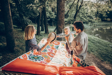 Young family playing with son on a hammock in park on camping trip, hipster mother smiling at baby...