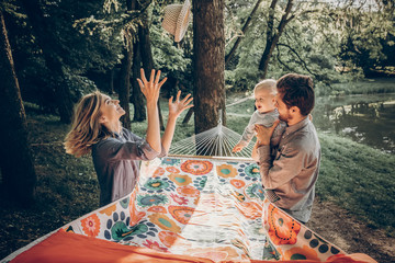 Young family playing with son on a hammock in park on camping trip, hipster mother putting a...