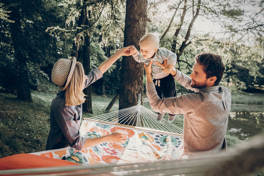 Young family playing with son on a hammock in park on camping trip, hipster mother smiling at baby boy near handsome father holding him, sunny morning background
