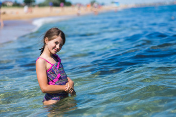 a little girl in a swimsuit stands waist-deep in clean sea water in front of the wave and looks at the camera