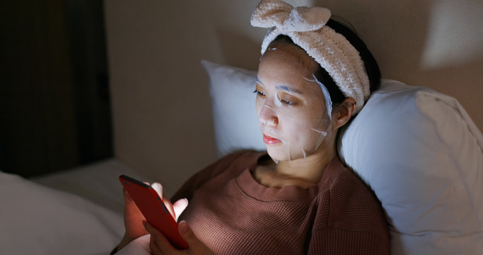 Woman uses mobile phone and applies face mask in bed at night