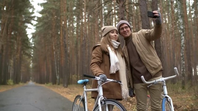 Romantic couple standing with bicycles on the road in park, kissing and looking at camera of smartphone while taking a selfie during ride on autumn day and then watching pictures