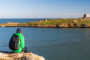 Tourist in a green jacket resting at the edge of a cliff and looking towards an island across the sea with a derelict church and a small defensive fort. Dalkey Island, Dublin, Ireland on a sunny day.