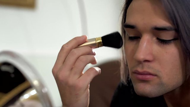 Portrait of handsome man doing makeup looking in the mirror close up. Transvestite man applies blush with makeup brush on his face. Concept of femininity in a man