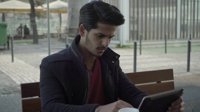 Serious young man using tablet computer on street. Handsome focused Indian man sitting on bench and working with digital tablet. Wireless technology concept