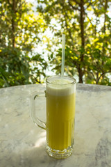 yellow fresh juice in a tall glass cup with a straw on a white marble table on a blurred background of green trees