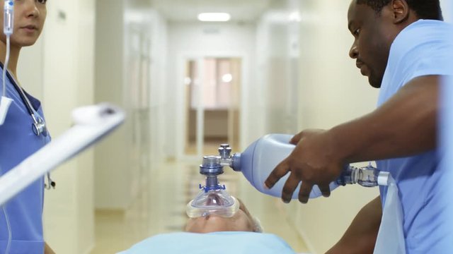 Handheld tracking shot of female ER nurse holding IV bag and talking to black male colleague administering oxygen to unresponsive patient while rushing him to surgery
