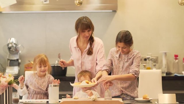 The mother puts a plate with just made biscuits on the table. The girls want to eat them right away, but their mum does not let them as the cookies are too hot to consume immediately.