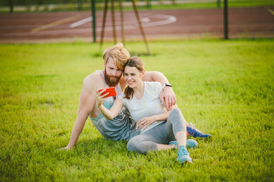 Sport and technology. Young in love heterosexual Caucasian couple resting after workout outdoors in park on lawn, green grass sitting in embrace and making selfie photo on smartphone camera at sunset