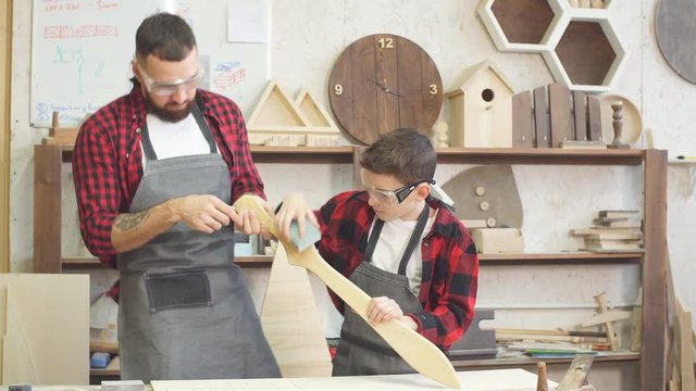 Little male woodworker creating his dream and his Dad is helping him. Close-up of young male carpenter examines with his son smoothness of DIY wooden sword to work with wood in his workshop