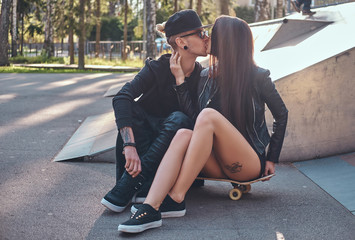 Fototapeta premium Young couple in love kissing while sitting on a skateboard and in a skate park at the summertime