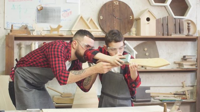 Fatherhood, hobby, carpentry, woodwork and people concept - Father and little son wearing protective glasses testing hand-made wood sword smooth surface at workshop