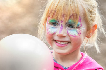 Glückliches geschminktes Mädchen beim Kinderkarneval auf einem Freizeitpark mit einem weißen Luftballon	