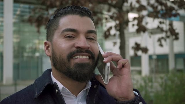 Cheerful bearded man talking by cell phone. Handsome happy man having pleasant phone talk and smiling at camera outdoor. Communication concept