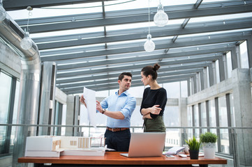 Two young architects with blueprints and model of a house standing in office, talking.