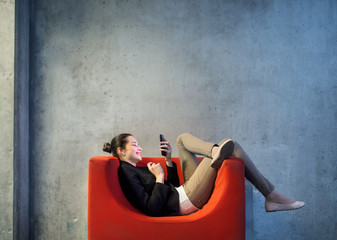 A young businesswoman with smartphone sitting on red armchair in office.