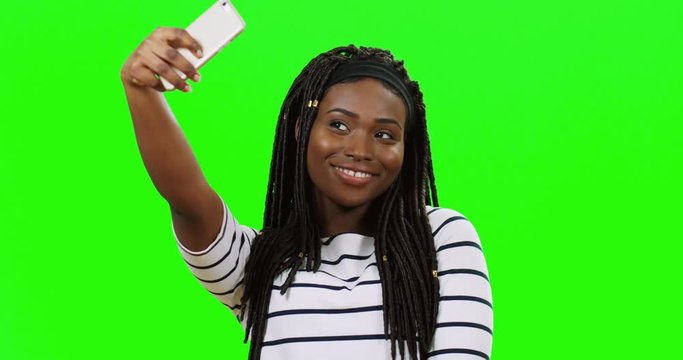 Charming young African American girl smiling and taking selfie photos on the smartphone camera while posing on the green screen background. Chroma key.