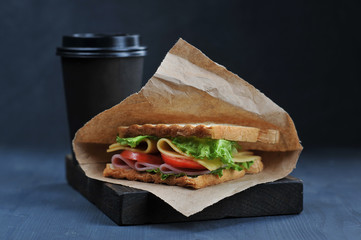 Sandwich with ham and cheese and paper cup with a drink on a dark background. Lettuce and tomato slices are used in the sandwich filling. The concept of fast food. Close-up. Macro shooting.