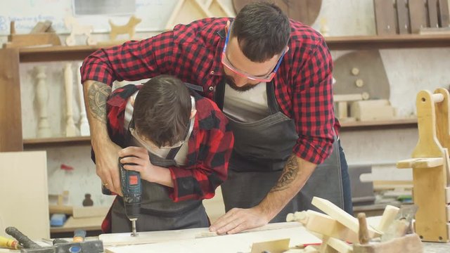Handsome father and his little pre-teen son dressed in checkered shirts and wearing protective glasses working with drill perforating wood plank at workshop