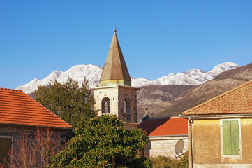 Fototapeta na wymiar Sunny winter day in Mediterranean village. Red tiled roofs and bell tower against blue sky and snow-capped mountain. Montenegro, Tivat, Donja Lastva village, Catholic Church of Saint Roch
